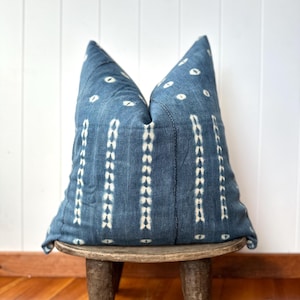 May include: A blue decorative pillow with a white dotted pattern and vertical stripes, resting on a small, round wooden stool. The pillow has a textured appearance and is filled, creating a soft, inviting look. The stool has three legs.
