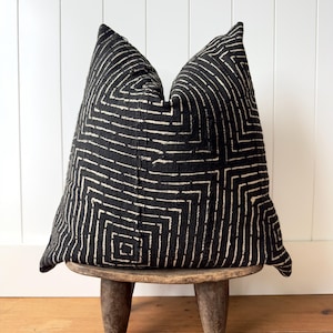 May include: A square throw pillow featuring a black fabric base with a white geometric pattern of concentric squares and lines. The pillow has a textured appearance and is displayed resting on a small, three-legged wooden stool with a dark finish.