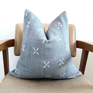 May include: A light blue decorative pillow with white cross-shaped patterns. The pillow is made of woven fabric and is placed on a beige chair with wooden arms. The pillow has a textured appearance and a soft, inviting look.