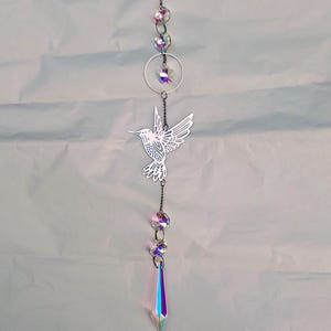 May include: A silver-colored hummingbird suncatcher with iridescent crystal accents. The suncatcher features a detailed hummingbird design, a circular ring, and several crystal beads. The bottom has a long, pointed crystal pendant.