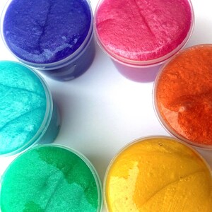 Puede incluir: Seis recipientes de slime de diferentes colores: azul, rosa, naranja, verde azulado, verde y amarillo. El slime es transparente y tiene una textura brillante.