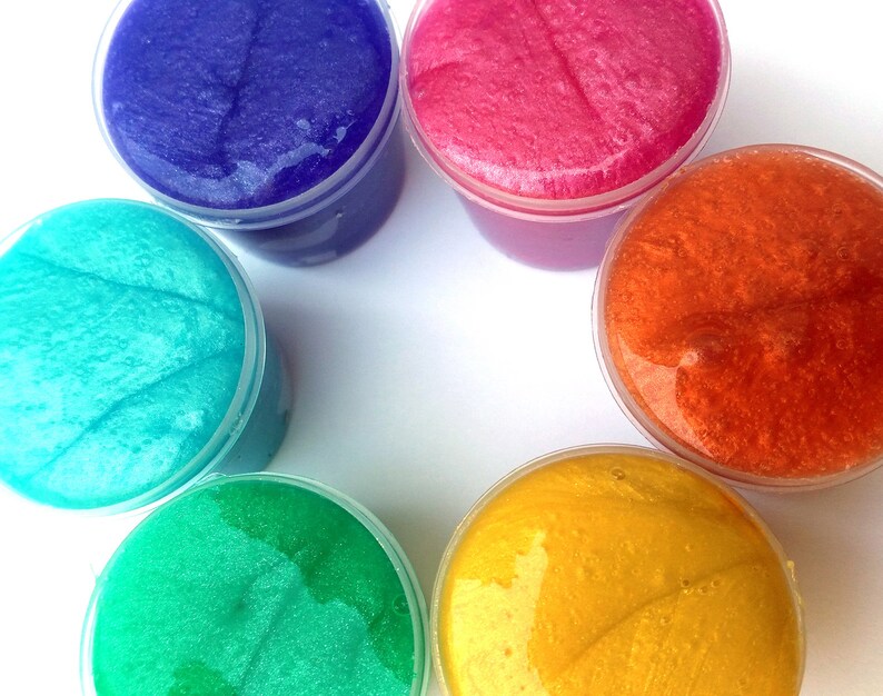 Puede incluir: Seis recipientes de slime de diferentes colores: azul, rosa, naranja, verde azulado, verde y amarillo. El slime es transparente y tiene una textura brillante.