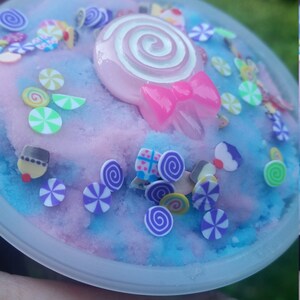Peut inclure: Un r&eacute;cipient en plastique transparent rempli de slime bleu et rose avec une vari&eacute;t&eacute; de paillettes color&eacute;es et une sucette rose et blanche sur le dessus.