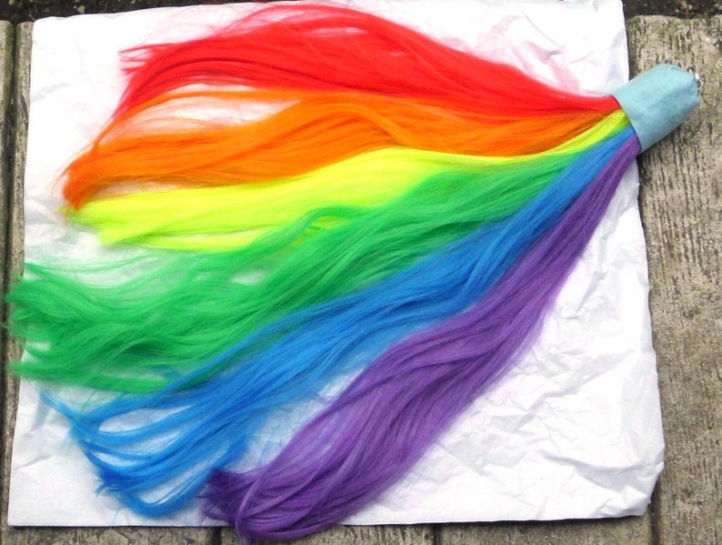 May include: A rainbow-colored hair extension with a blue fabric band at the end. The hair is arranged in a cascade of colors, starting with red at the top and ending with purple at the bottom.