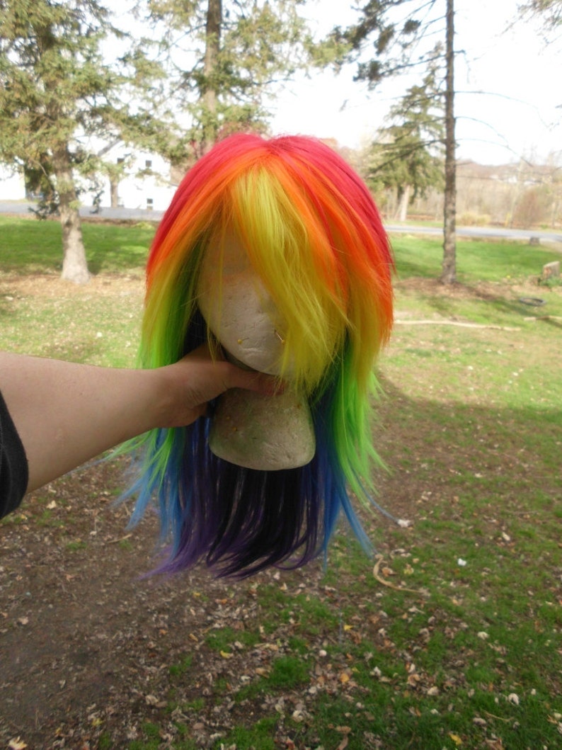 May include: A rainbow colored wig with long, straight hair. The wig is on a styrofoam head and is being held by a hand.