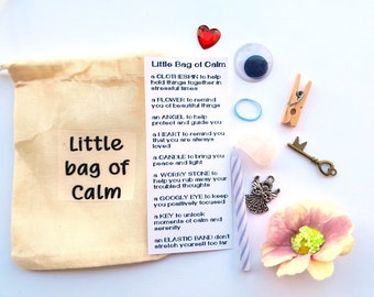 May include: A small, beige cloth bag with the words "Little bag of Calm" printed on it. The bag contains a variety of small items, including a red heart, a blue googly eye, a wooden clothespin, a blue rubber band, a small key, a pink and white flower, a silver angel charm, a white candle, and a pink worry stone. The bag is a gift for someone who needs a little peace and quiet.