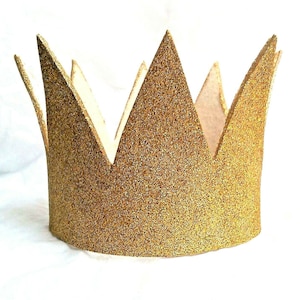 May include: A gold glitter crown with a pointed shape. The crown is made of cardboard and has a simple design.