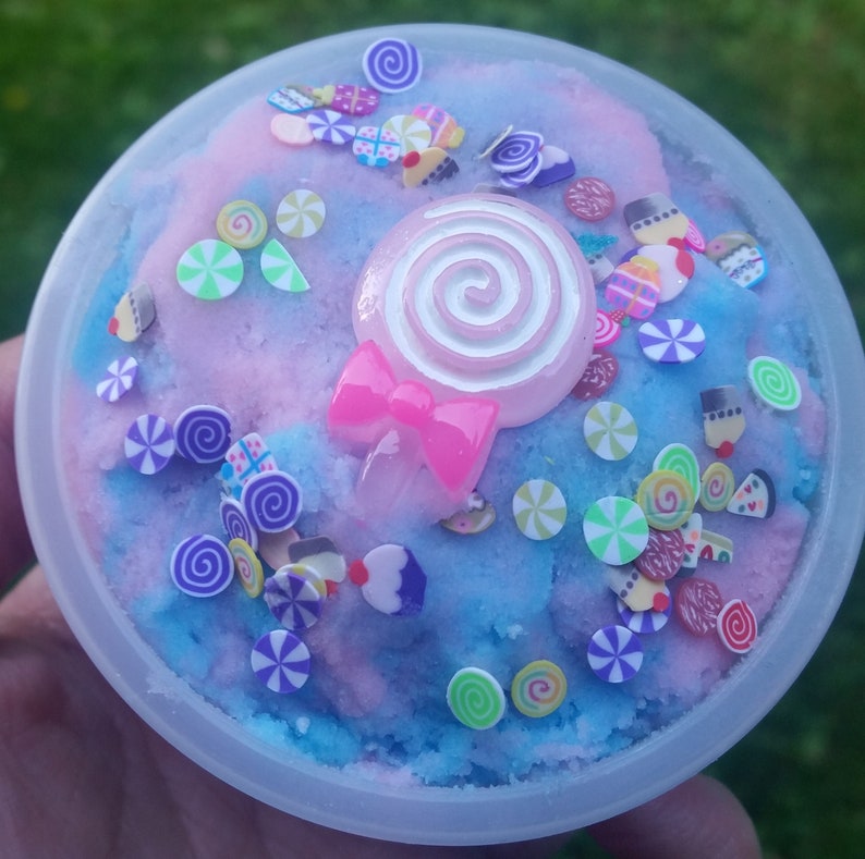 Peut inclure: Un r&eacute;cipient en plastique transparent rempli de slime bleu et rose avec des paillettes color&eacute;es et une sucette rose avec un tourbillon blanc sur le dessus.