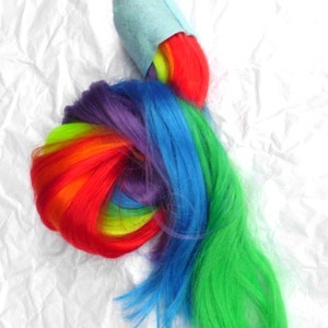 May include: A rainbow-colored ponytail extension with a light blue fabric band at the top. The extension is made of synthetic hair and is tied in a knot.