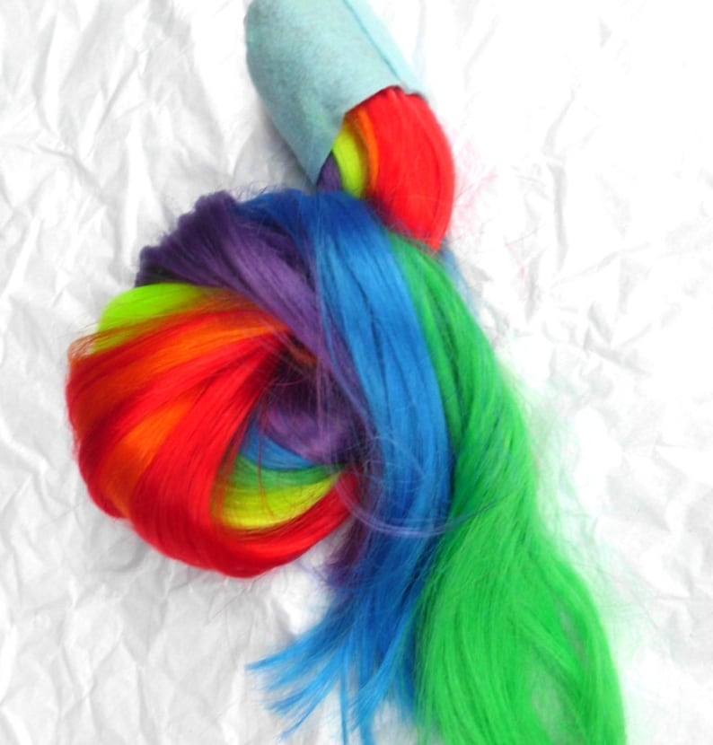 May include: A rainbow-colored ponytail extension with a light blue fabric band at the top. The extension is made of synthetic hair and is tied in a knot.
