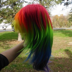 May include: A long, straight, rainbow-colored wig with red, yellow, green, blue, and purple streaks. The wig is styled with long layers and is being held by a hand.