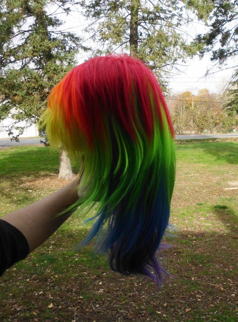 May include: A long, straight, rainbow-colored wig with red, yellow, green, blue, and purple streaks. The wig is styled with long layers and is being held by a hand.