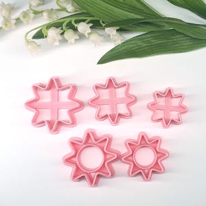 May include: A collection of five pink, star-shaped cookie cutters in varying sizes. The cutters have a sunburst design with a cross in the center. The image also includes white flowers and green leaves in the background.