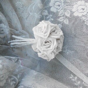 May include: A white fabric rose bouquet with a stem made of white fabric. The bouquet is lying on a white lace fabric.