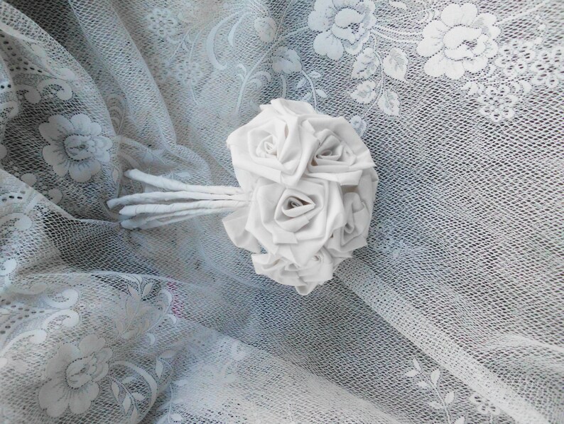 May include: A white fabric rose bouquet with a stem made of white fabric. The bouquet is lying on a white lace fabric.