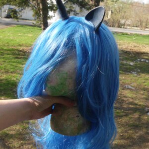 May include: A blue wig with a black unicorn horn and ears. The wig is on a mannequin head.