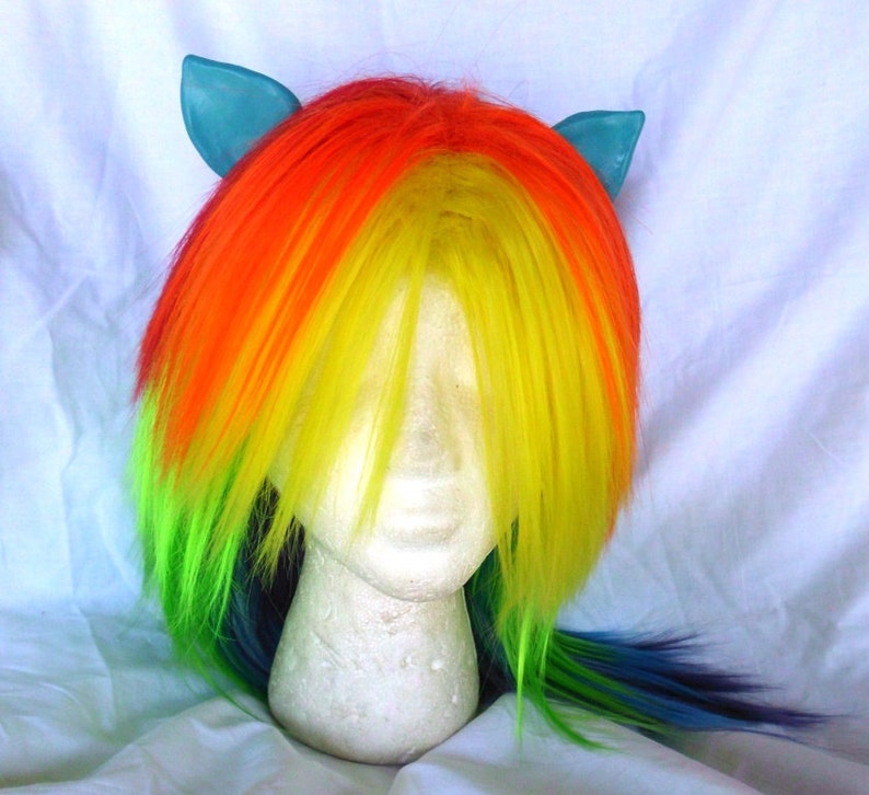 May include: A rainbow-colored wig with blue ears. The wig is styled with long, straight hair and a side part. The wig is on a white mannequin head.