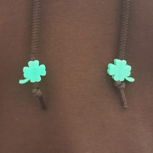 May include: Two dark brown braided drawstrings hanging against a matching brown fabric background. Each drawstring features a bright mint green four-leaf clover charm secured by a small knot at the base of the clover, providing a decorative accent.
