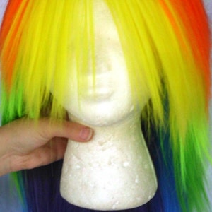 May include: A rainbow-colored wig with blue ear clips. The wig is made of synthetic hair and is styled in a long, straight fashion. The wig is on a white mannequin head.