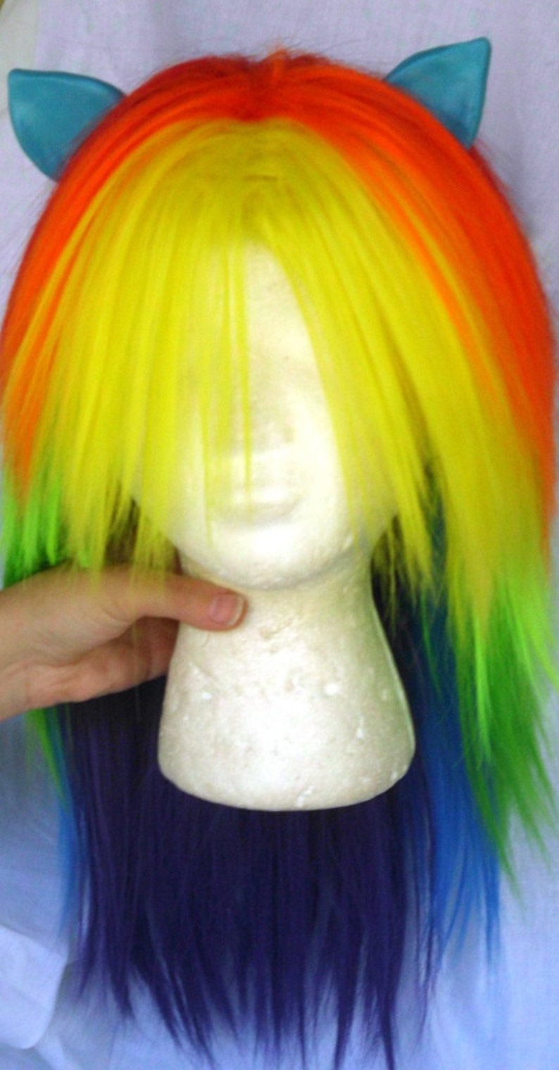 May include: A rainbow-colored wig with blue ear clips. The wig is made of synthetic hair and is styled in a long, straight fashion. The wig is on a white mannequin head.