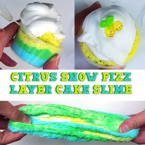 Puede incluir: Slime de pastel en capas Citrus Snow Fizz en un vaso de pl&aacute;stico transparente con una cobertura de crema batida blanca. El slime est&aacute; en capas de azul, verde y amarillo. El slime se est&aacute; sosteniendo en una mano.