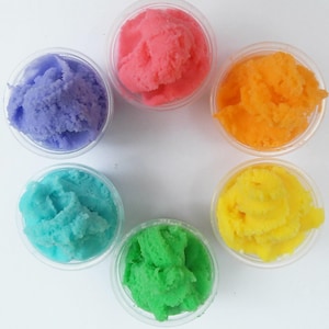 Puede incluir: Seis pequeños recipientes transparentes llenos de glaseado batido esponjoso y colorido. Los colores son morado, rosa, naranja, amarillo, azul y verde.