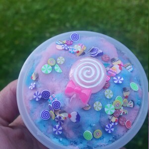 Peut inclure: Un r&eacute;cipient en plastique transparent rempli de slime bleu et rose avec une vari&eacute;t&eacute; de paillettes color&eacute;es et une sucette rose avec un tourbillon blanc sur le dessus.