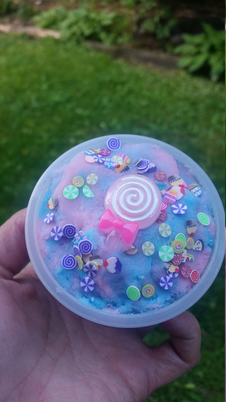 Peut inclure: Un r&eacute;cipient en plastique transparent rempli de slime bleu et rose avec une vari&eacute;t&eacute; de paillettes color&eacute;es et une sucette rose avec un tourbillon blanc sur le dessus.