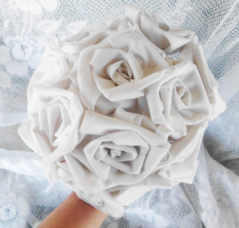May include: A bouquet of white fabric roses, arranged in a circular shape. The roses are made from a soft, white fabric and have a realistic appearance.