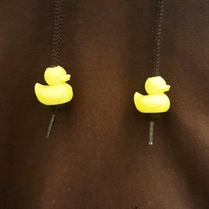 May include: A dark brown hooded garment featuring two black drawstring cords. Each cord is finished with a small, bright yellow rubber duck bead acting as an aglet. The ducks are oriented to face outward, providing a distinct decorative detail.