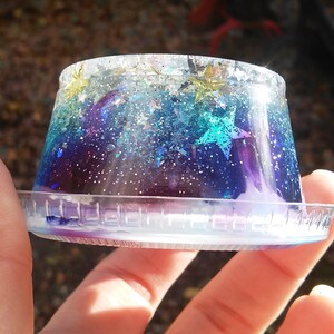 Puede incluir: Un recipiente de pl&aacute;stico transparente lleno de resina de color galaxia azul, p&uacute;rpura y verde con purpurina y estrellas doradas.