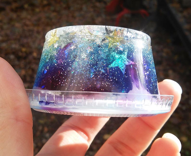 Puede incluir: Un recipiente de pl&aacute;stico transparente lleno de resina de color galaxia azul, p&uacute;rpura y verde con purpurina y estrellas doradas.