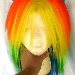 May include: A rainbow colored wig with blue ear-shaped hair clips. The wig is styled in a long, straight, layered style. The wig is on a mannequin head.