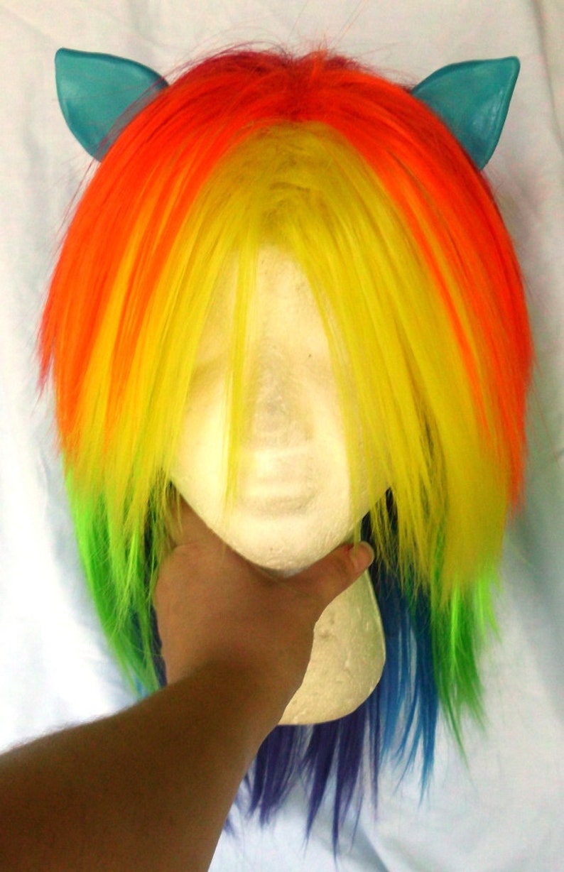 May include: A rainbow colored wig with blue ear-shaped hair clips. The wig is styled in a long, straight, layered style. The wig is on a mannequin head.