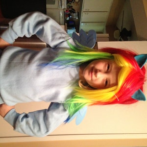May include: A young girl wearing a blue sweatshirt and a rainbow-colored wig with blue ears. The wig has streaks of red, yellow, blue, and green.