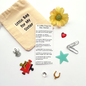 May include: A small white fabric bag with a tag that reads "A Little Bag for My Sister". The tag has a poem about sisterhood with a list of small items, including paperclips, an angel charm, a puzzle piece, a flower, a diamond, a star, a horseshoe, and a heart.