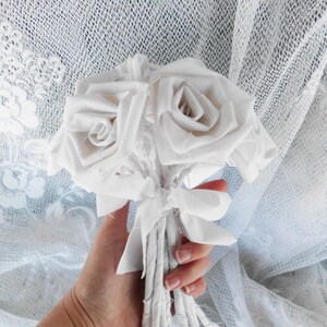 May include: A bouquet of white fabric roses tied together with white fabric ribbons. The bouquet is held in a hand against a white lace background.