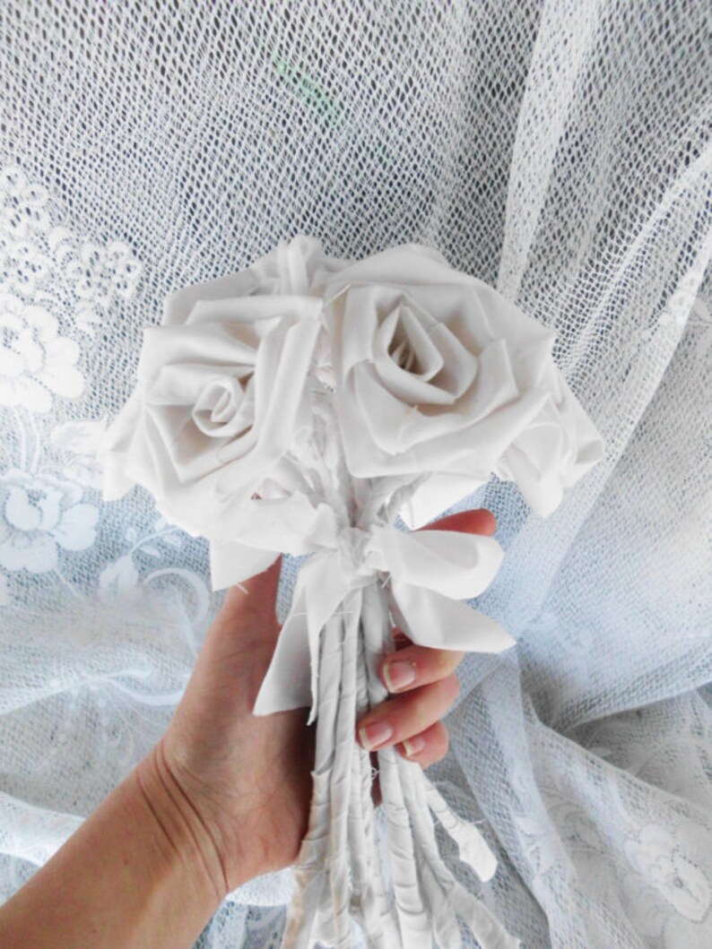 May include: A bouquet of white fabric roses tied together with white fabric ribbons. The bouquet is held in a hand against a white lace background.