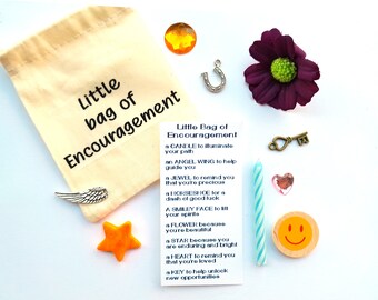 May include: A small white canvas bag with the words "Little bag of Encouragement" printed on it. The bag is filled with small trinkets, including a candle, a horseshoe, a key, a star, a flower, a heart, a smiley face, and a silver angel wing. The bag also includes a card with the words "Little Bag of Encouragement" and a list of items with their meanings.