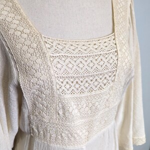 May include: Cream-colored dress with a square neckline and long sleeves. The bodice features intricate lace detailing around the neckline and upper chest. The dress has a flowing, lightweight fabric.