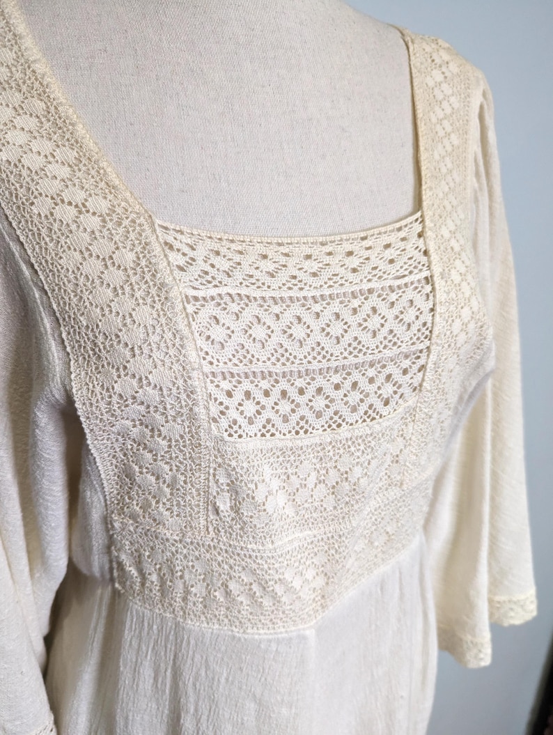 May include: Cream-colored dress with a square neckline and long sleeves. The bodice features intricate lace detailing around the neckline and upper chest. The dress has a flowing, lightweight fabric.