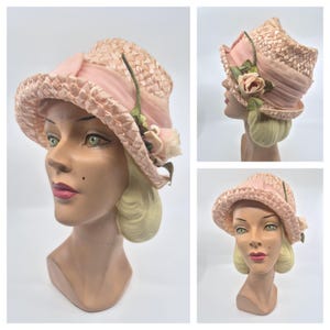 May include: A light pink woven straw hat featuring a wide, pleated satin ribbon band around the crown. The hat is decorated with a single fabric rose in a matching pale pink hue, accented by two green fabric leaves and a thin green stem.