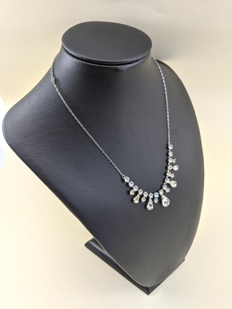 Puede incluir: Un collar de cadena de plata con un colgante de cristal en forma de l&aacute;grima. El colgante est&aacute; rodeado de cristales m&aacute;s peque&ntilde;os, creando un efecto en cascada.