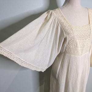 May include: Off-white vintage dress with wide sleeves and a square neckline. The dress features delicate lace trim along the neckline and sleeve edges. The fabric has a subtle textured appearance, giving it a soft, flowing look.