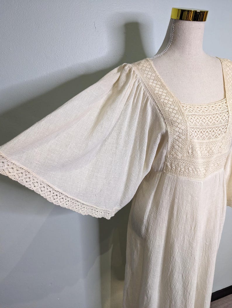 May include: Off-white vintage dress with wide sleeves and a square neckline. The dress features delicate lace trim along the neckline and sleeve edges. The fabric has a subtle textured appearance, giving it a soft, flowing look.