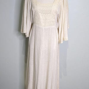 May include: Cream-colored vintage dress with a square neckline and lace detailing on the bodice and sleeves. The dress has a flowing, floor-length skirt and is displayed on a mannequin with a gold base.