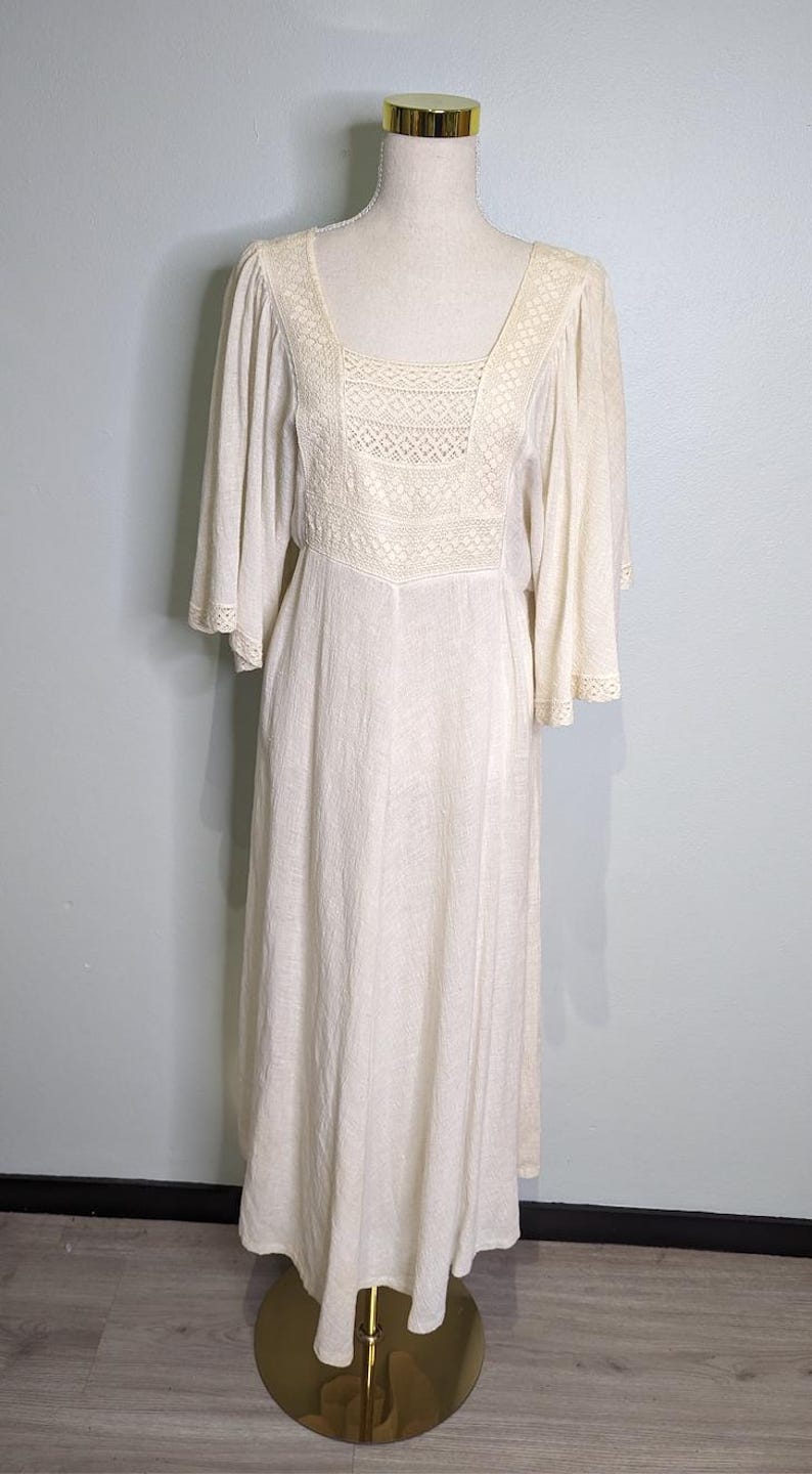 May include: Cream-colored vintage dress with a square neckline and lace detailing on the bodice and sleeves. The dress has a flowing, floor-length skirt and is displayed on a mannequin with a gold base.