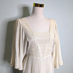 May include: Cream-colored vintage dress with a square neckline and three-quarter sleeves. The bodice features intricate lace detailing, and the sleeves and hem are trimmed with delicate lace. The dress is displayed on a mannequin against a light blue background.