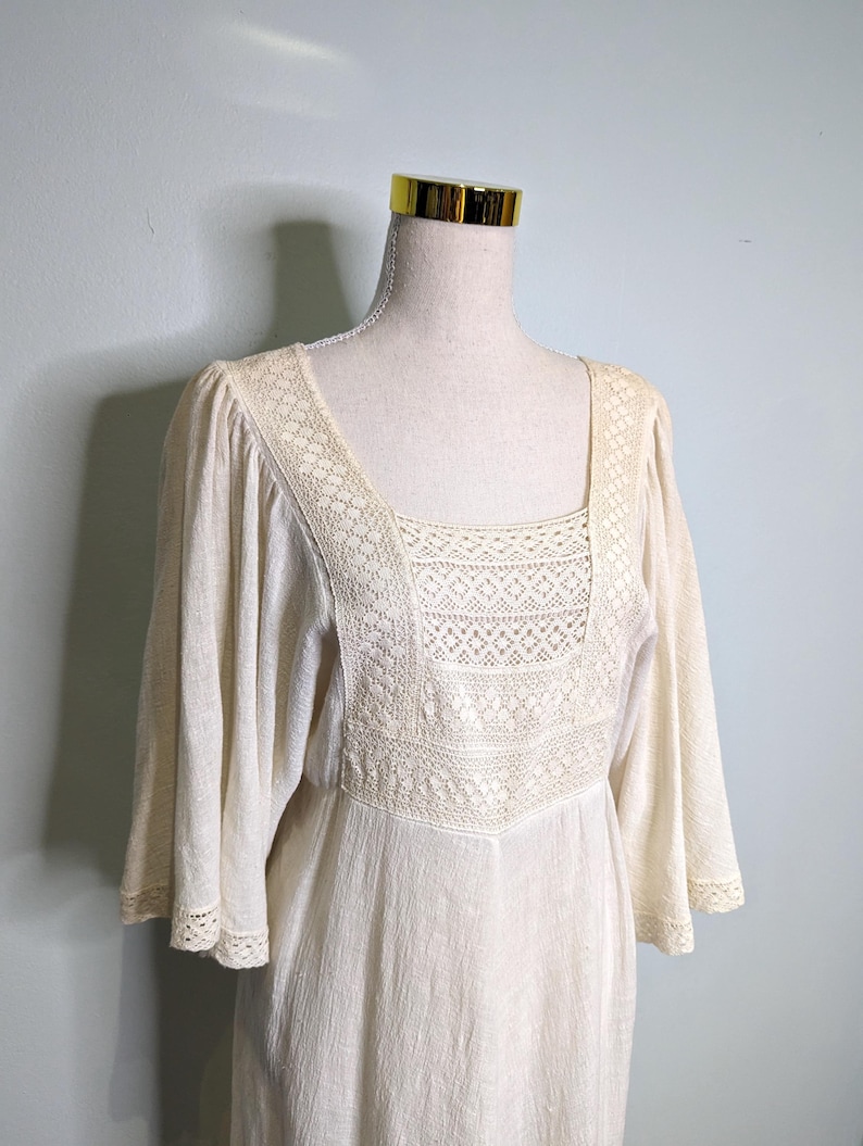May include: Cream-colored vintage dress with a square neckline and three-quarter sleeves. The bodice features intricate lace detailing, and the sleeves and hem are trimmed with delicate lace. The dress is displayed on a mannequin against a light blue background.