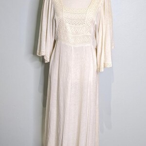 May include: A cream-colored vintage dress with a square neckline and bell sleeves. The bodice features a detailed lace panel. The dress is displayed on a mannequin with a gold base, set against a light blue wall and wooden floor.